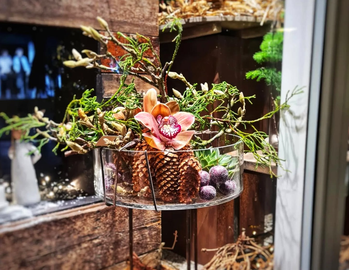 Glasfad arrangeret med kogler, orkidéer og grene. 